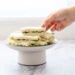 A hand reaching to pick up a cucumber sandwich from a white cake stand of sandwiches.