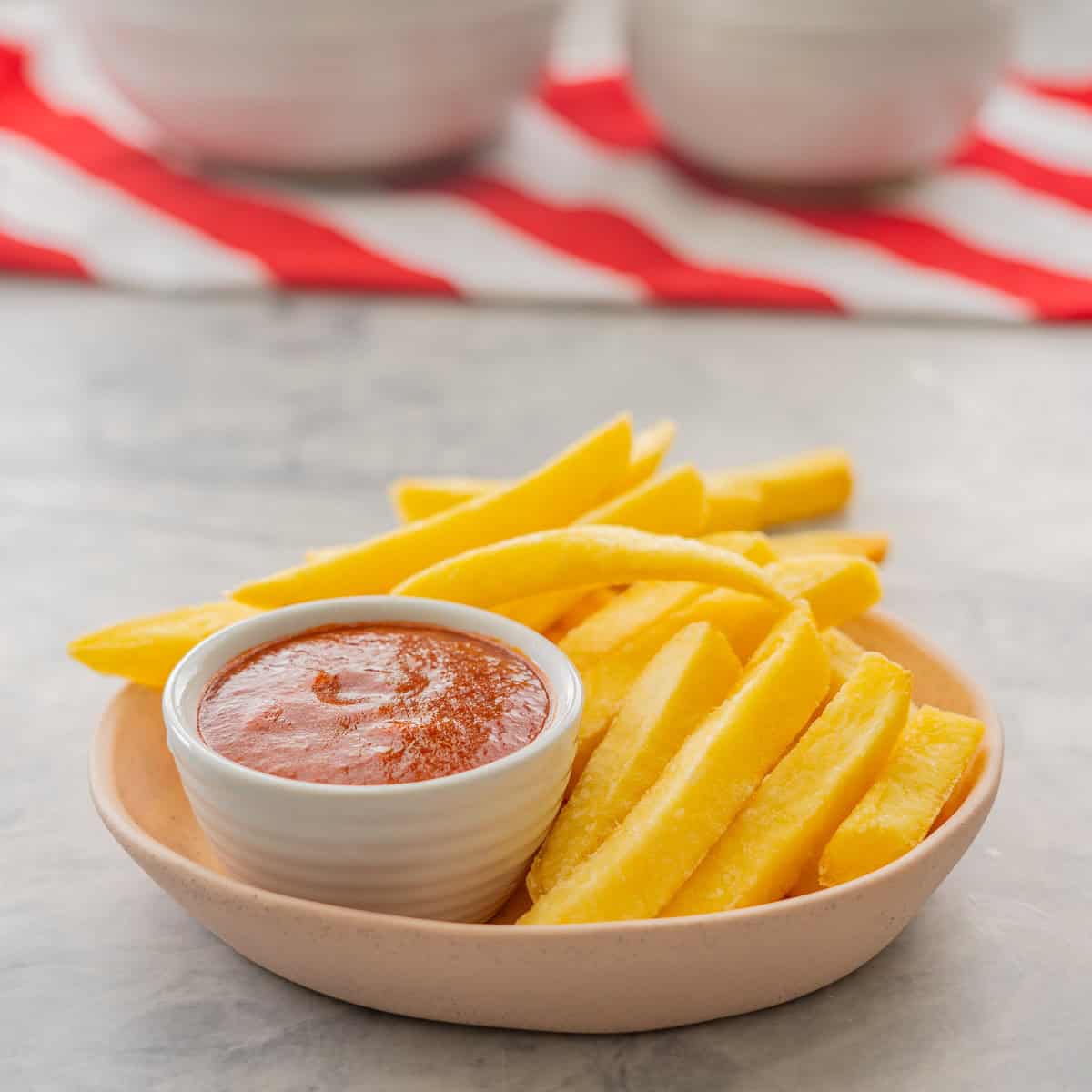 A bowl of fries with homemade ketchup in small ramekin inside bowl on benchtop with folded red and white stripped table cloth.