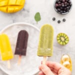 A hand holding a kiwifruit popsicle above a plate which has one each of a mango and blueberry popsicle resting on it. The bench surrounding the plate has got sliced mango, kiwifruit, blueberries and bananas scattered around it.