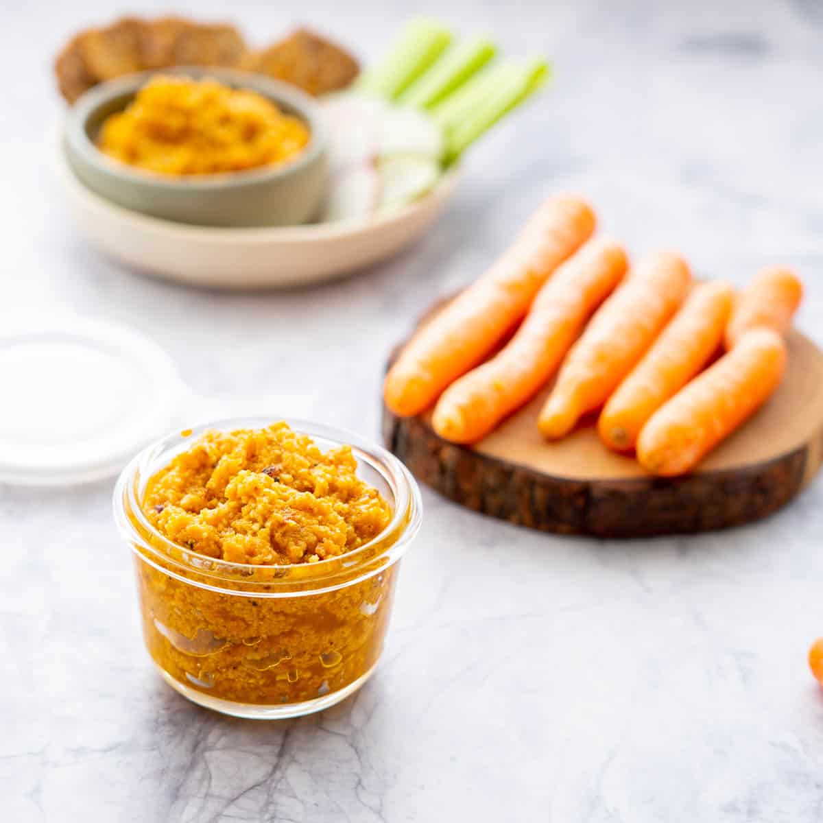 A small glass jar filled with orange pesto, on a bench with a snack platter and loose carrots.