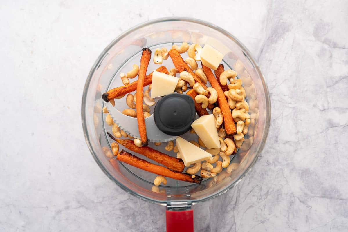 A food processor with roasted carrots, chunks of parmesan and cashew nuts. 
