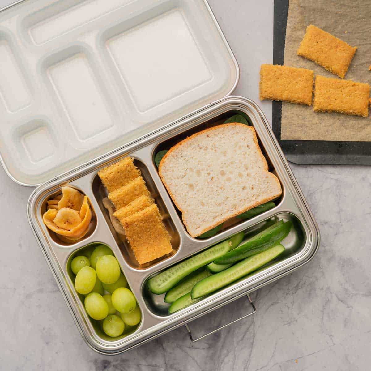 A stainless steel bento box packed with a sandwich, crackers, fruit and vegetables.