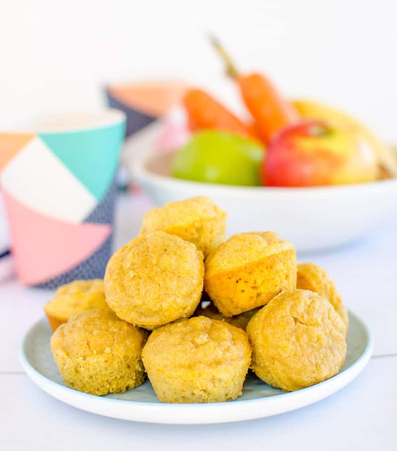 A plate of 10 mini muffins, with a bowl of apples, carrots and bananas in the background.