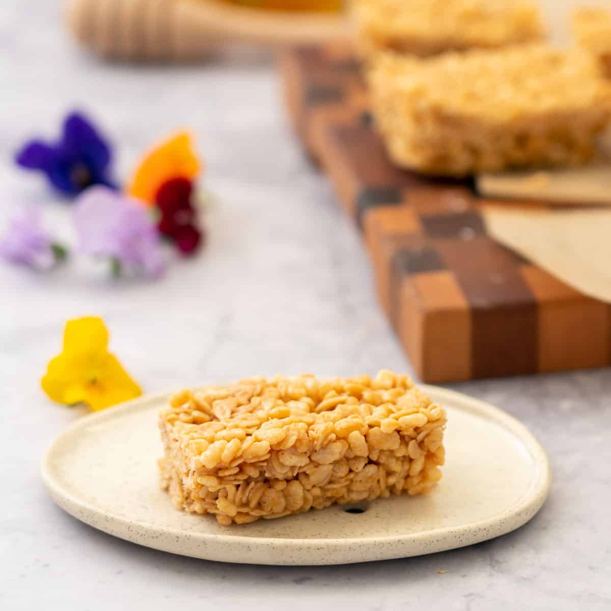 A piece of peanut butter rice krispie slice on a side plate next to a handful of edible flowers.