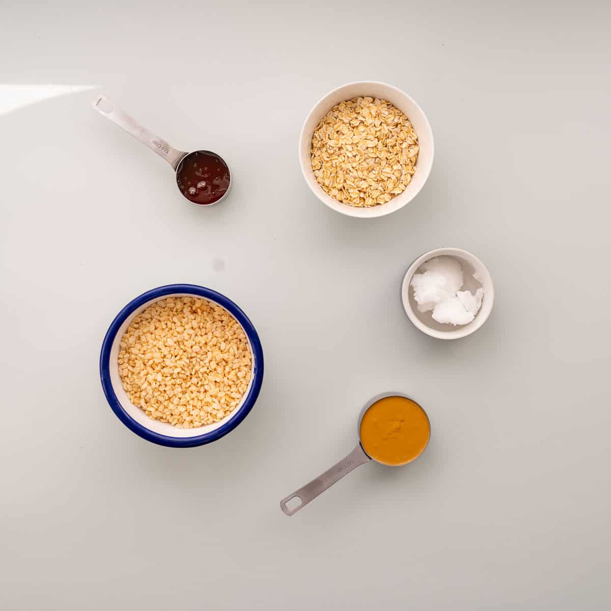 Honey, coconut oil, peanut butter, rice bubbles and rolled oats in bowls and measuring cups on a bench top.