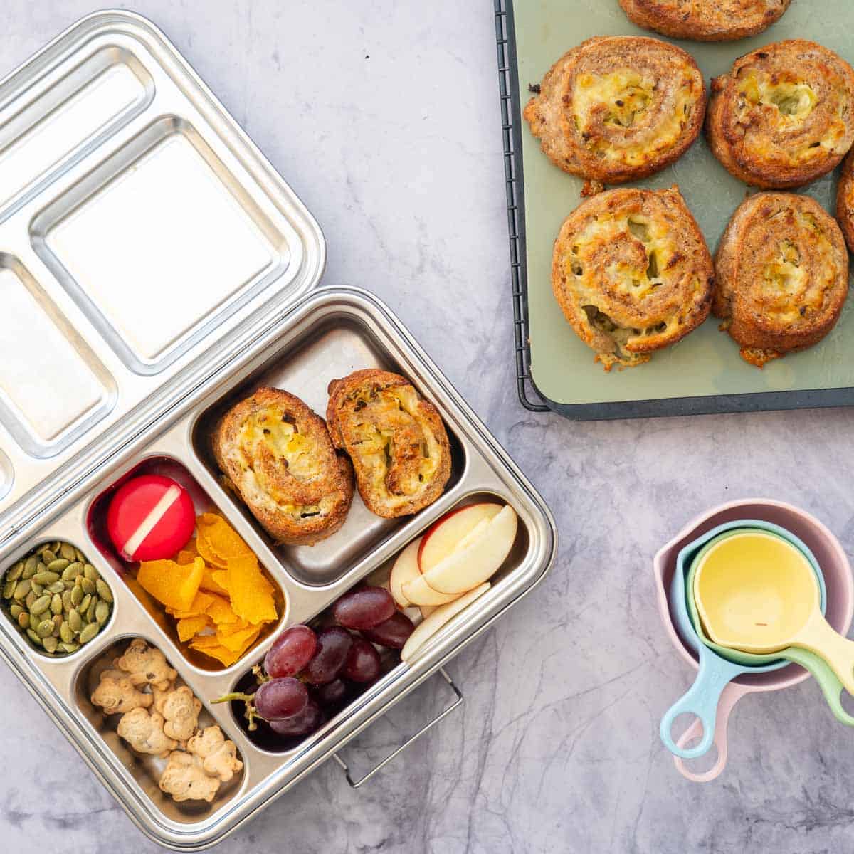 A stainless steel lunch box packed with leek scrolls, fruit, vegetables, cheese and crackers.