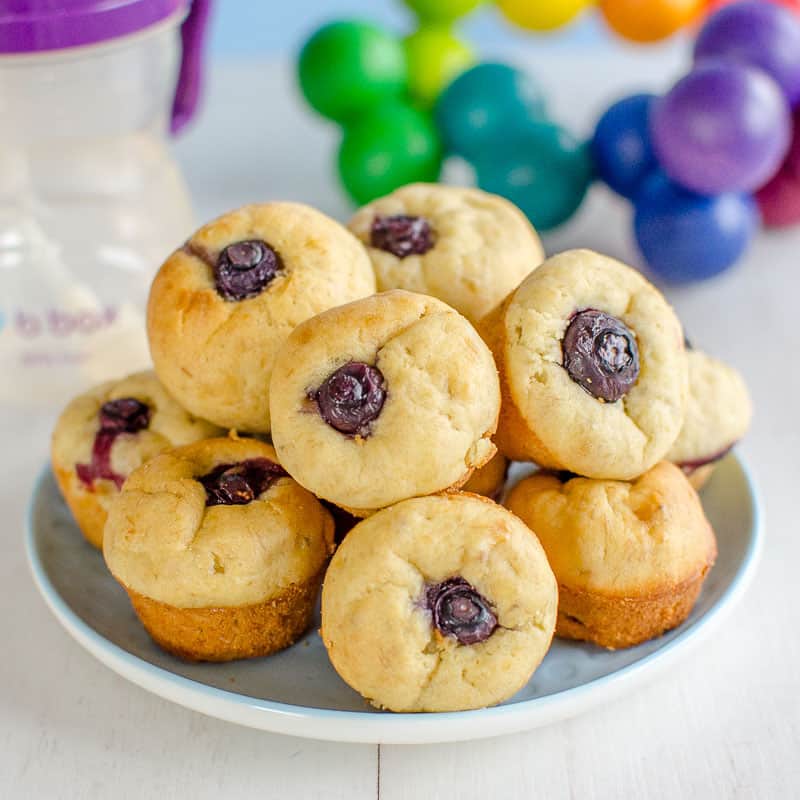 A plate of mini blueberry banana muffins, a baby sippy cup and wooden toy in the background.