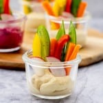 A small glass jar with hummus n the bottom and batons of colourful vegetables standing up in the cup