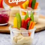 A small glass jar with hummus n the bottom and batons of colourful vegetables standing up in the cup with text overlay for pinterest