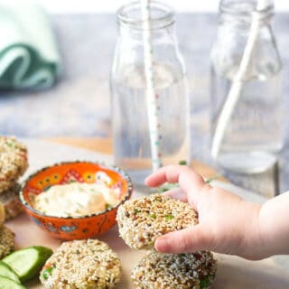 Three tuna quinoa cakes  on a chopping board with dipping sauce, childs hand grabbing a quinoa cake.