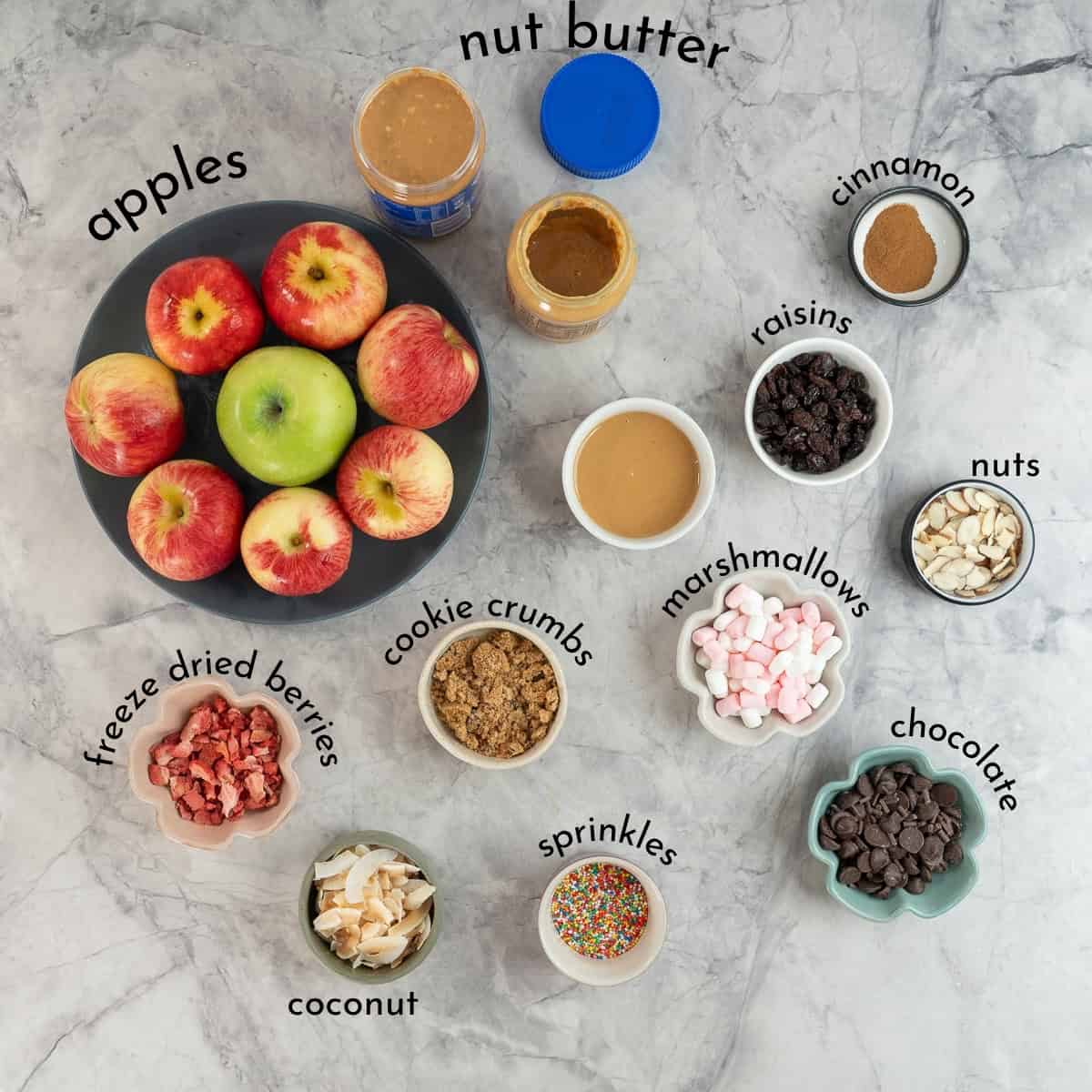 Ingredients for apple donuts on benchtop with text overlay, apples, nut butter, sprinkles, dried fruits, chocolate chips, cocoaut etc. 