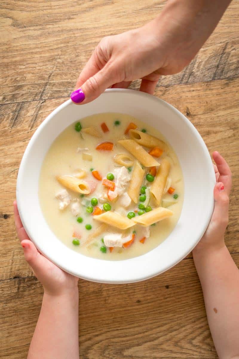 creamy chicken pasta soup, adult hands with purple finger nails handing bowl of soup to child