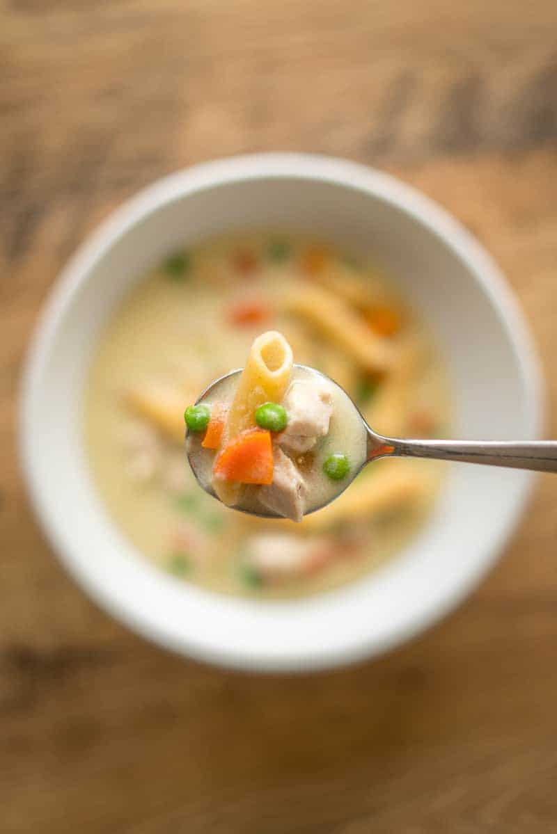 creamy chicken pasta soup with pasta, close up image of a soup in a spoon above a white bowl