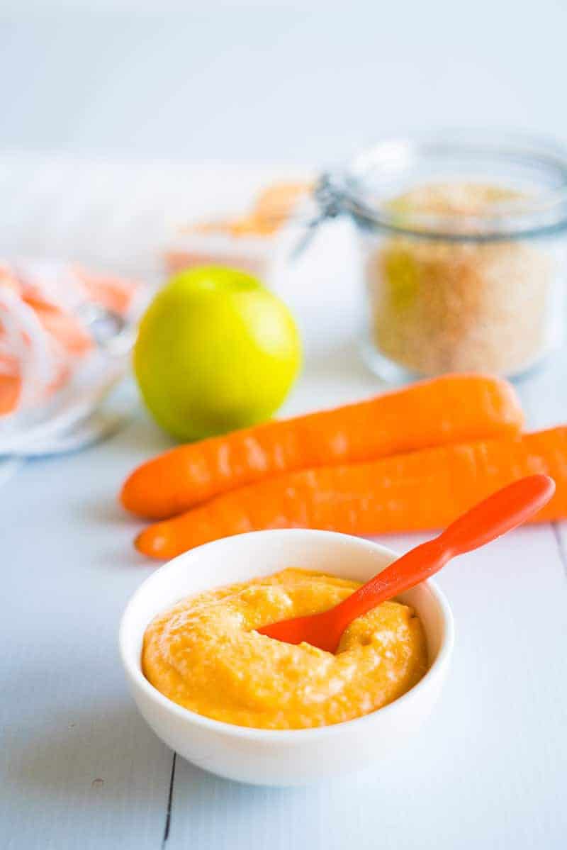 homemade quinoa baby cereal in foreground in a white bowl with carrot and apples in the background