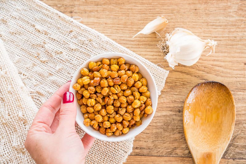 Crunchy garlic roasted chickpeas, in a small white bowl on a linen surface, hand holding the bowl
