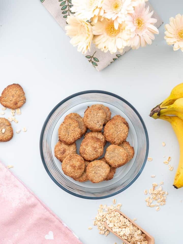 A round glass container full of oatmeal cookies on a bench next to a bunch of bananas.
