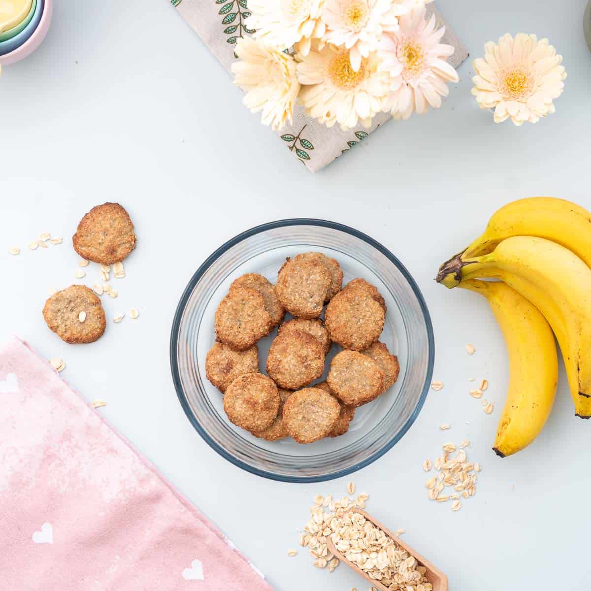 A round glass container full of oatmeal cookies on a bench next to a bunch of bananas.