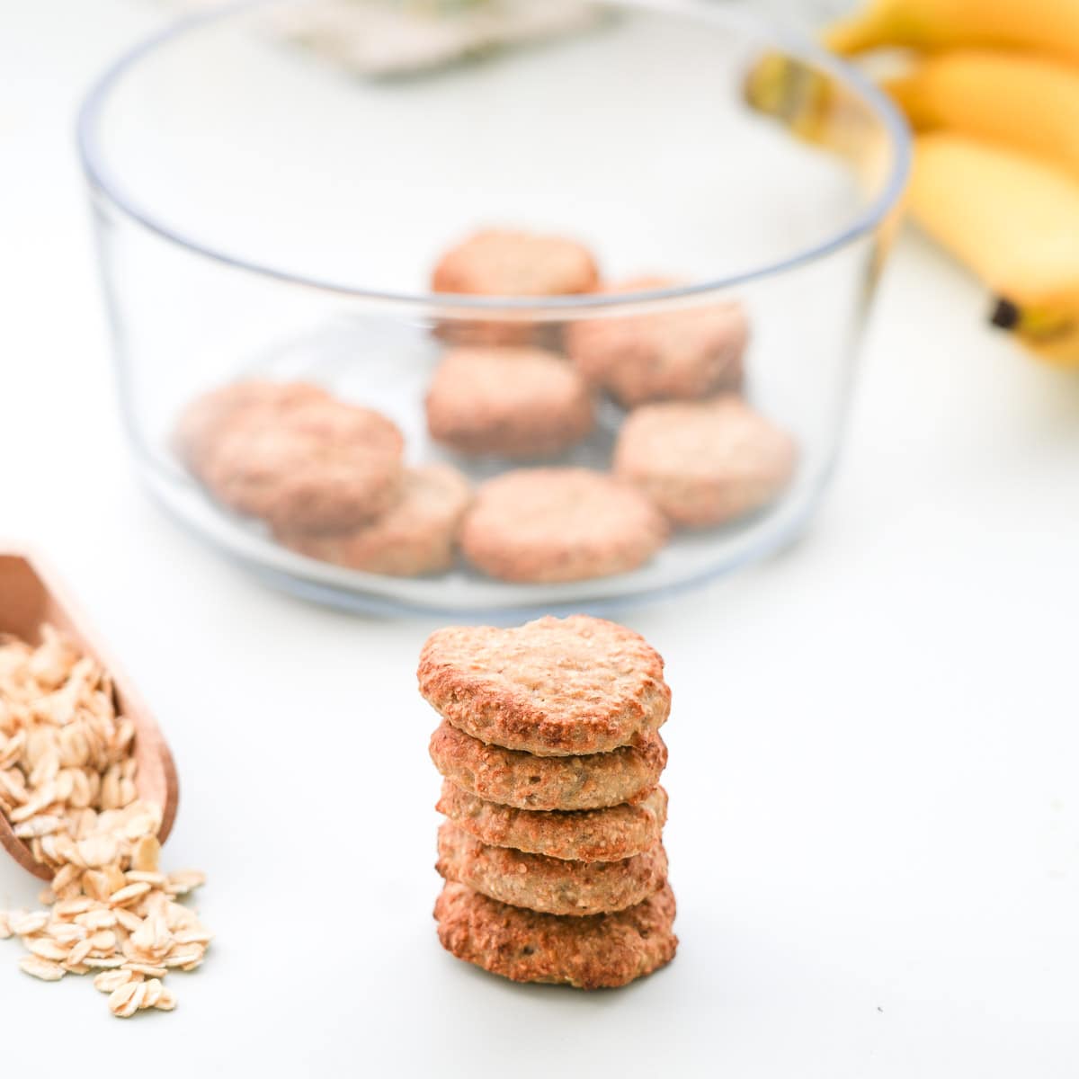 A tower of five small oatmeal cookies on a bench in-front of a container of cookies.