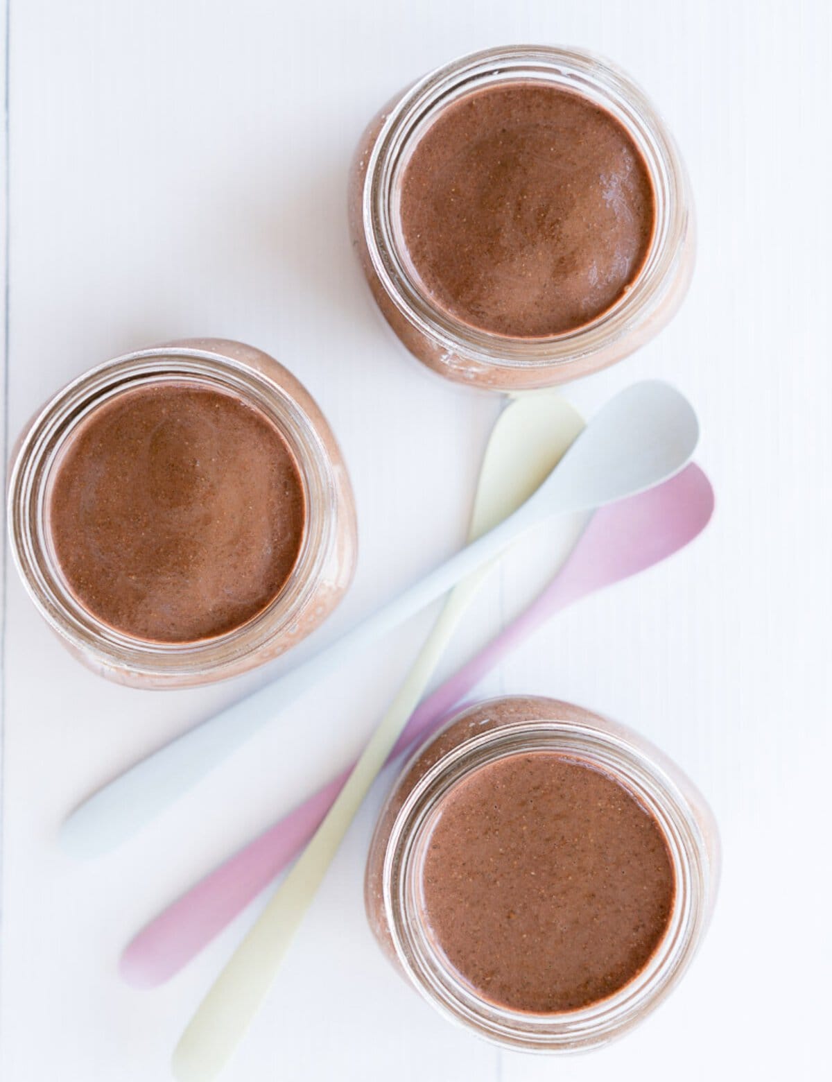 Looking down into three glass jars filled with smooth chocolate pudding next to three long handled dessert spoons. 