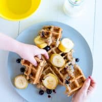 waffles on a plate, with banana and blueberries, 2 children's hands reaching for the waffles