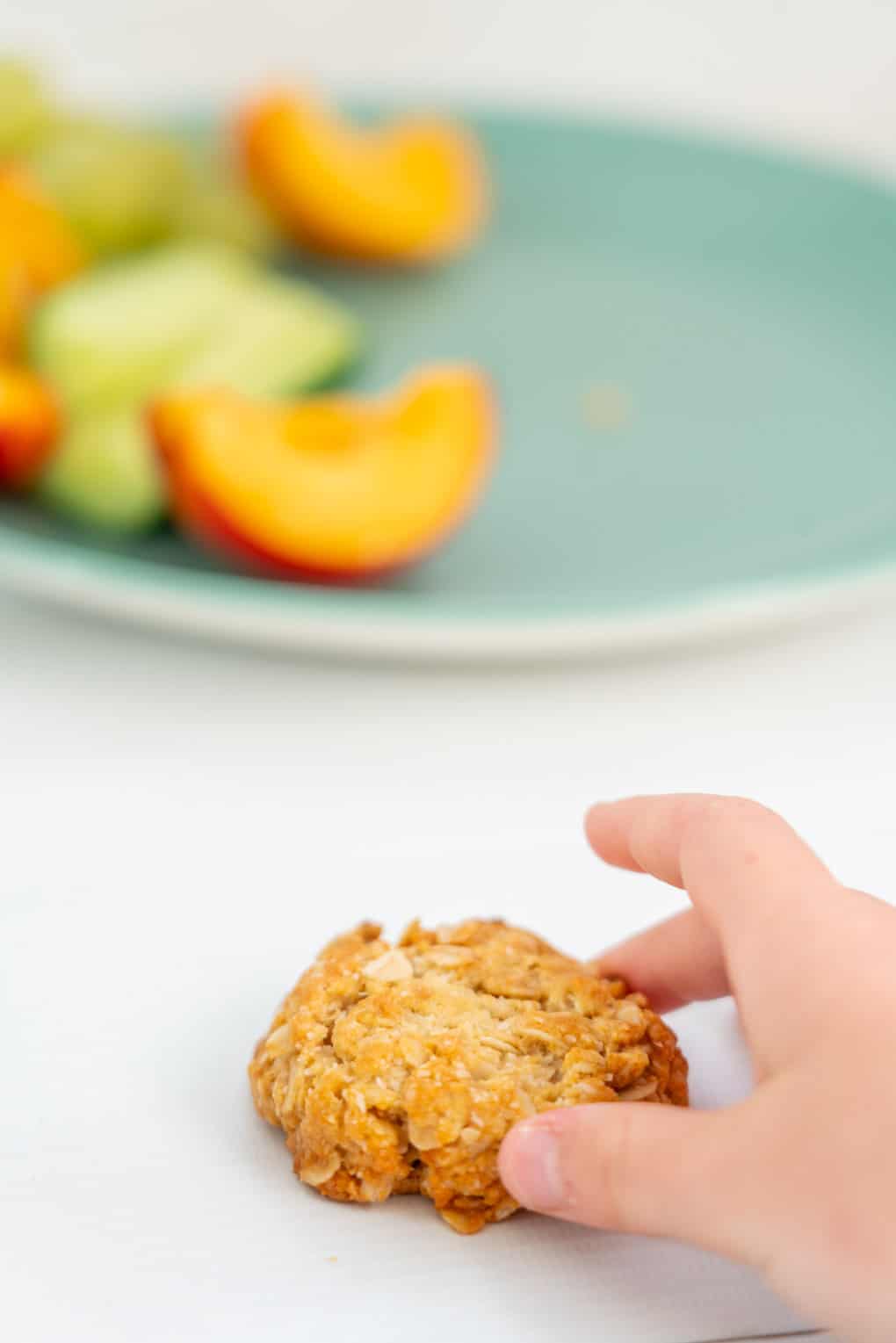 A child's hand reaching for a golden oat cookie on a white bench top
