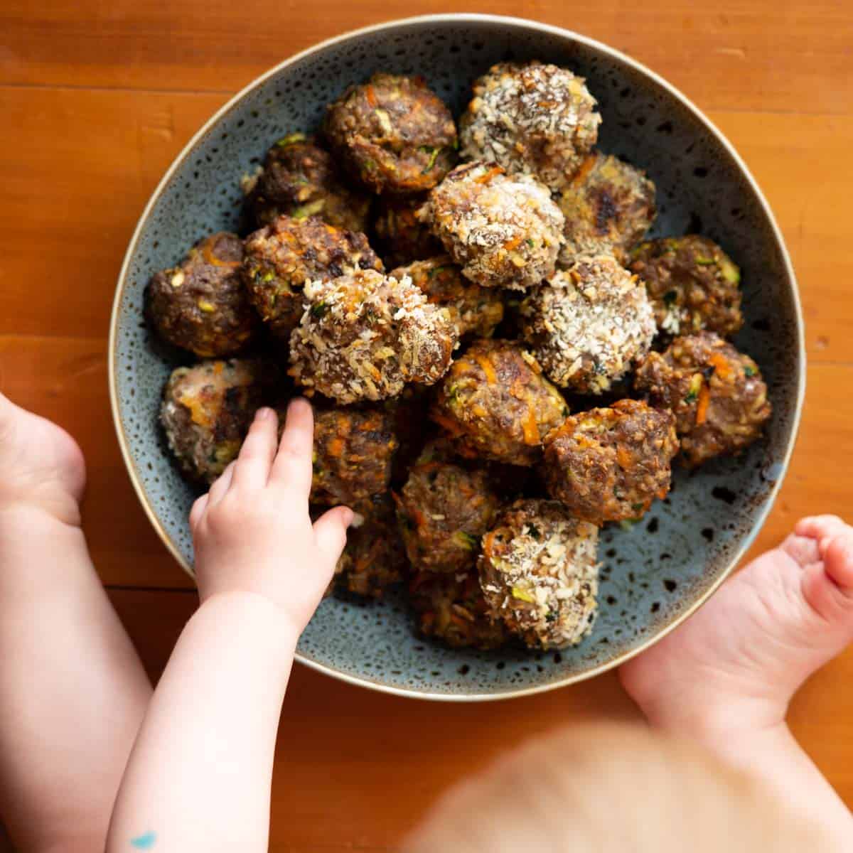 A large bowl of rissoles sitting on a wooden floor, a baby's hand reaching for one. 