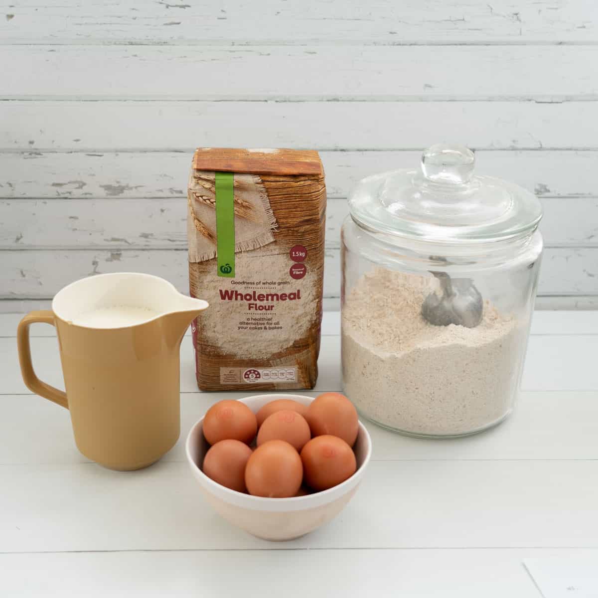 jug of milk, bowl of eggs, bag of wholemeal flour and large glass flour jar with scoop