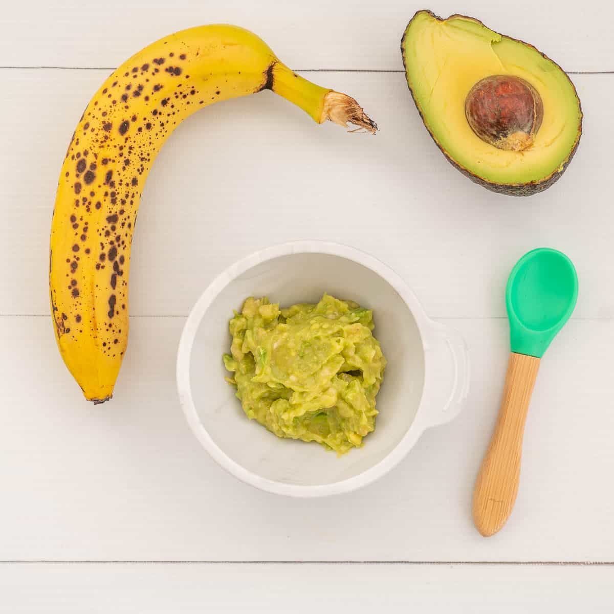 mashed avocado and banana baby food in a white baby bowl with green spoon