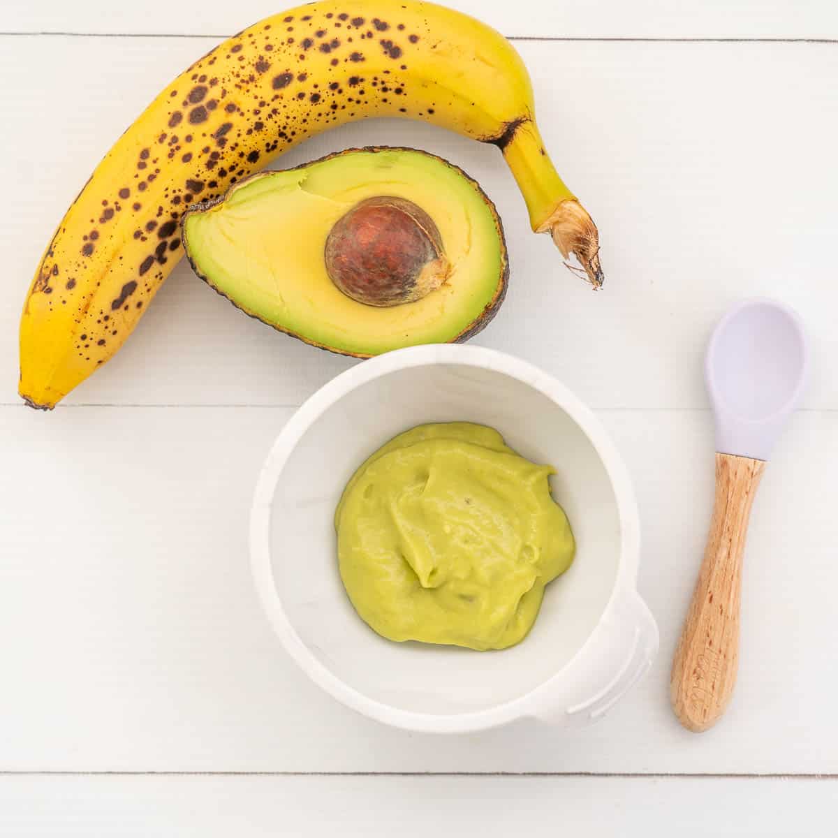 smooth green avocado baby food in a white baby bowl next to a banana and avocado