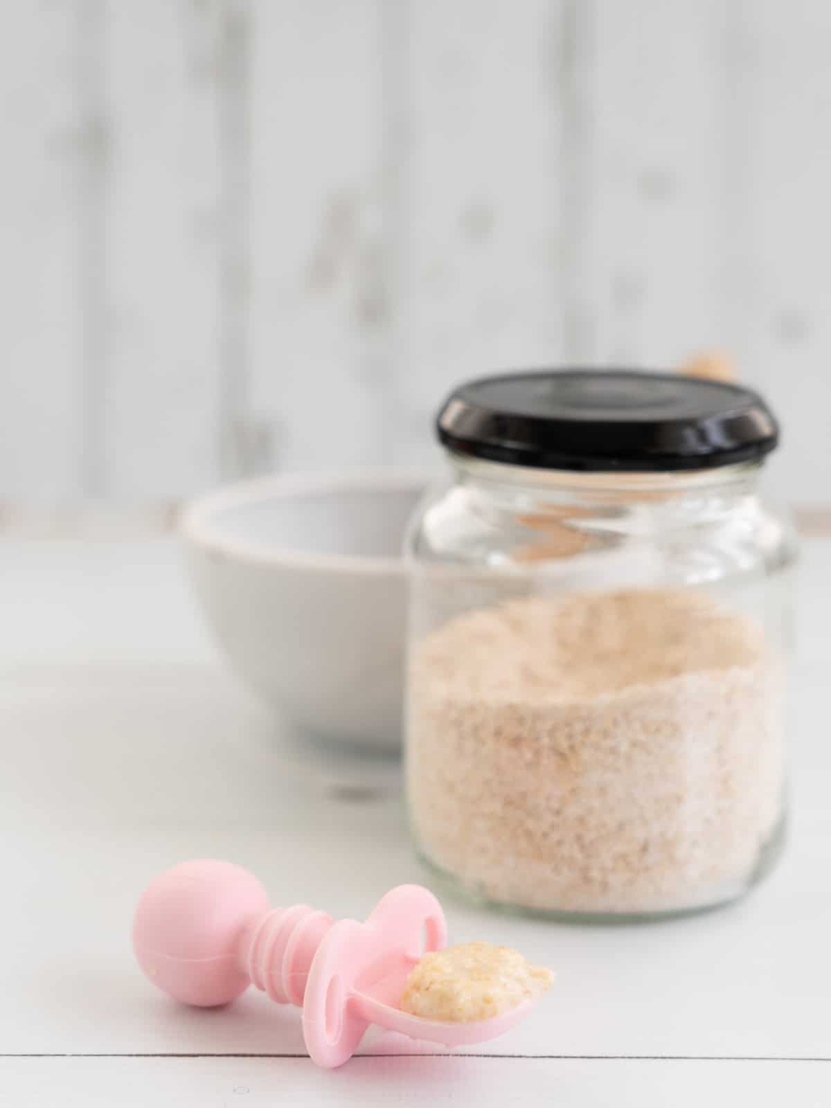 spoonful of cooked baby porridge on a pink baby spoon with a jar of baby oats in the background