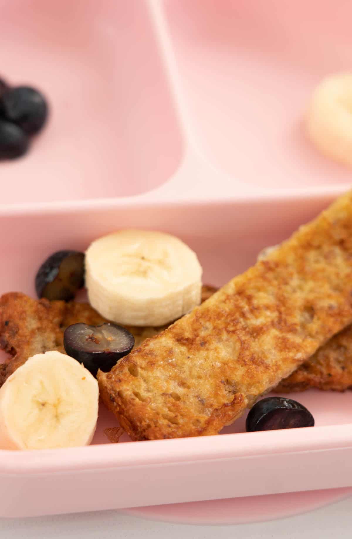 a pink baby breakfast plate with eggy bread, blueberries and sliced bananas