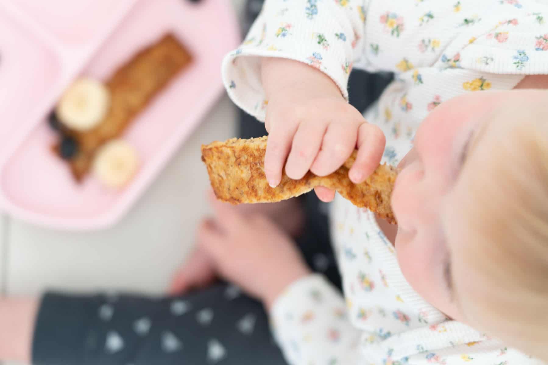 A baby eating a piece of eggy bread while seated on the floor next to a pink baby plate. 