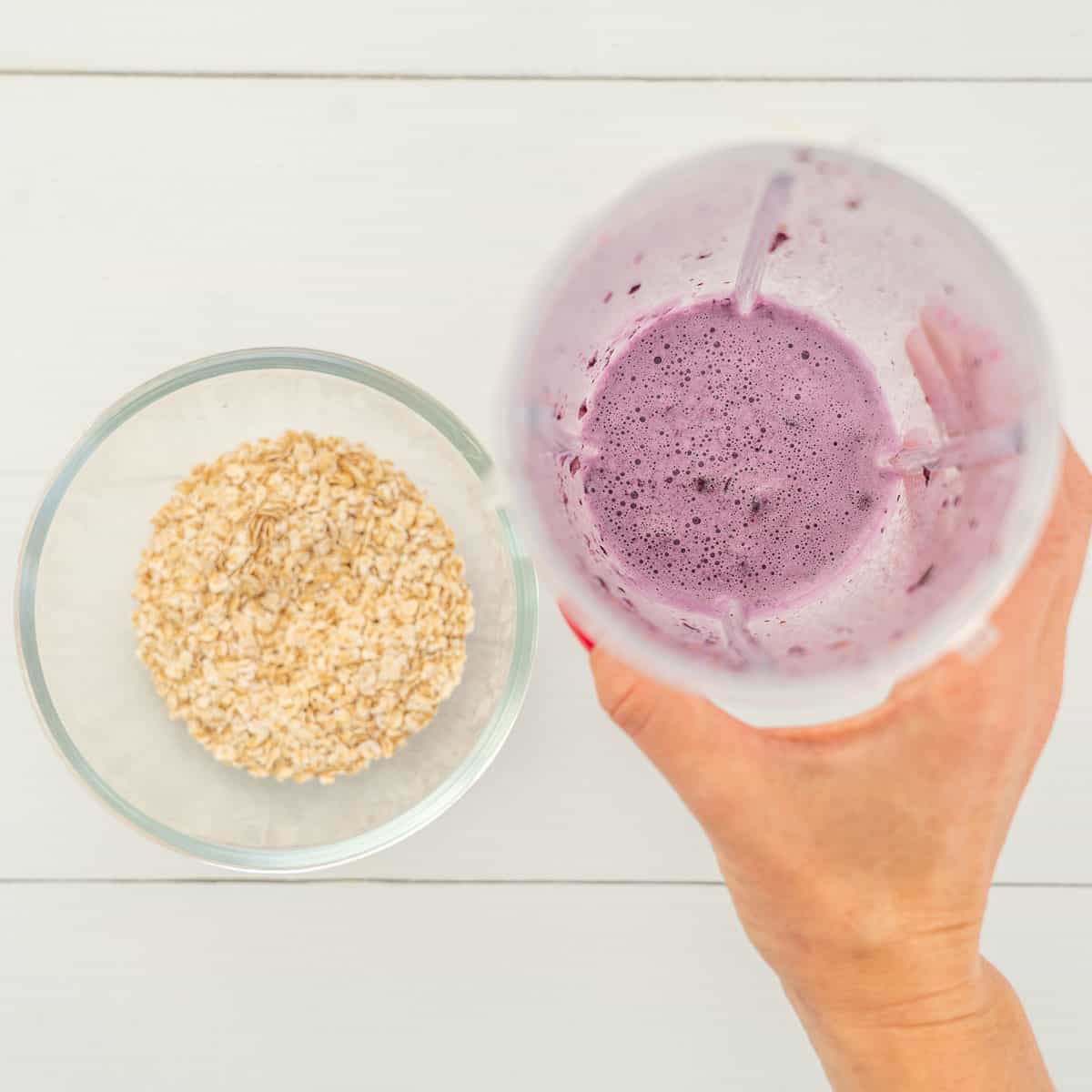 Hand holding a smoothie cup with blueberry mixture inside and oats in a glass bowl on bench top.