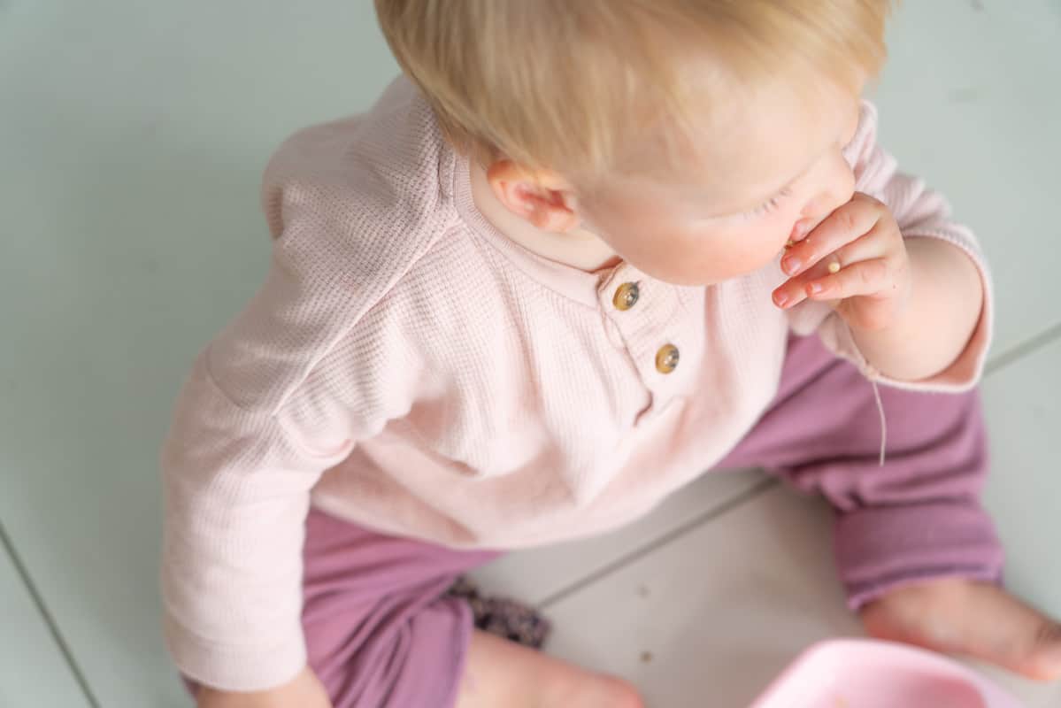 Baby dressed in a pink top and purple pants eating a porridge finger