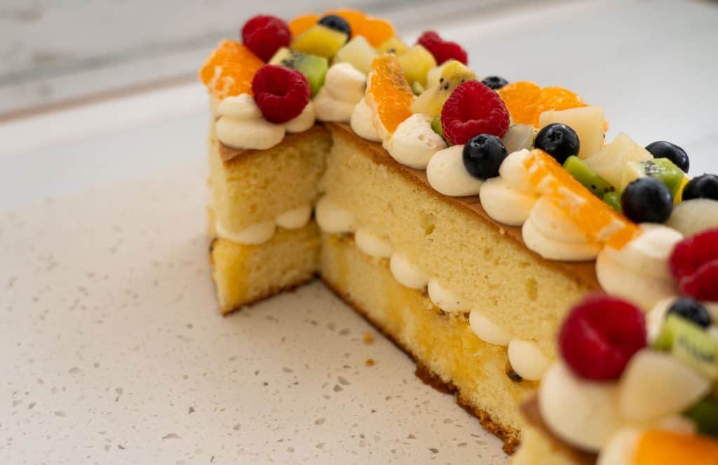 two layers of sponge cakes with piped whipped cream in the middle. The top of the cake is decorated with whipped cream and fruit