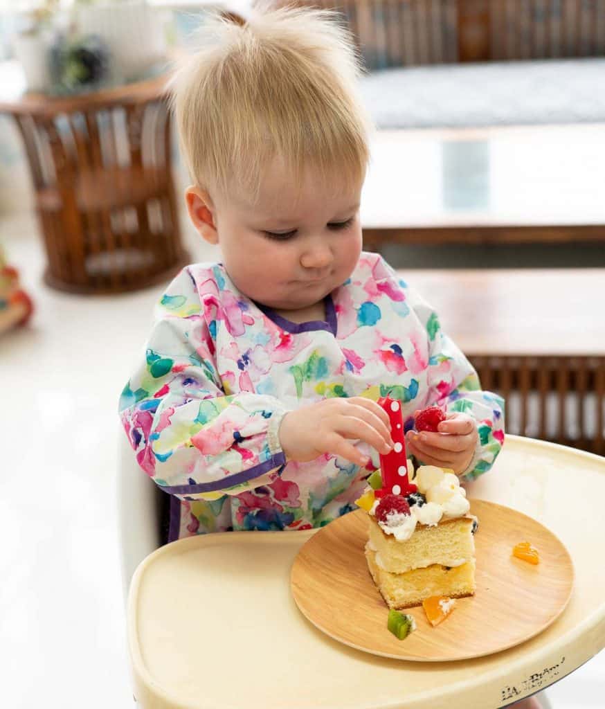 a baby in a high chair with a square fruit covered cake and a red number one candle