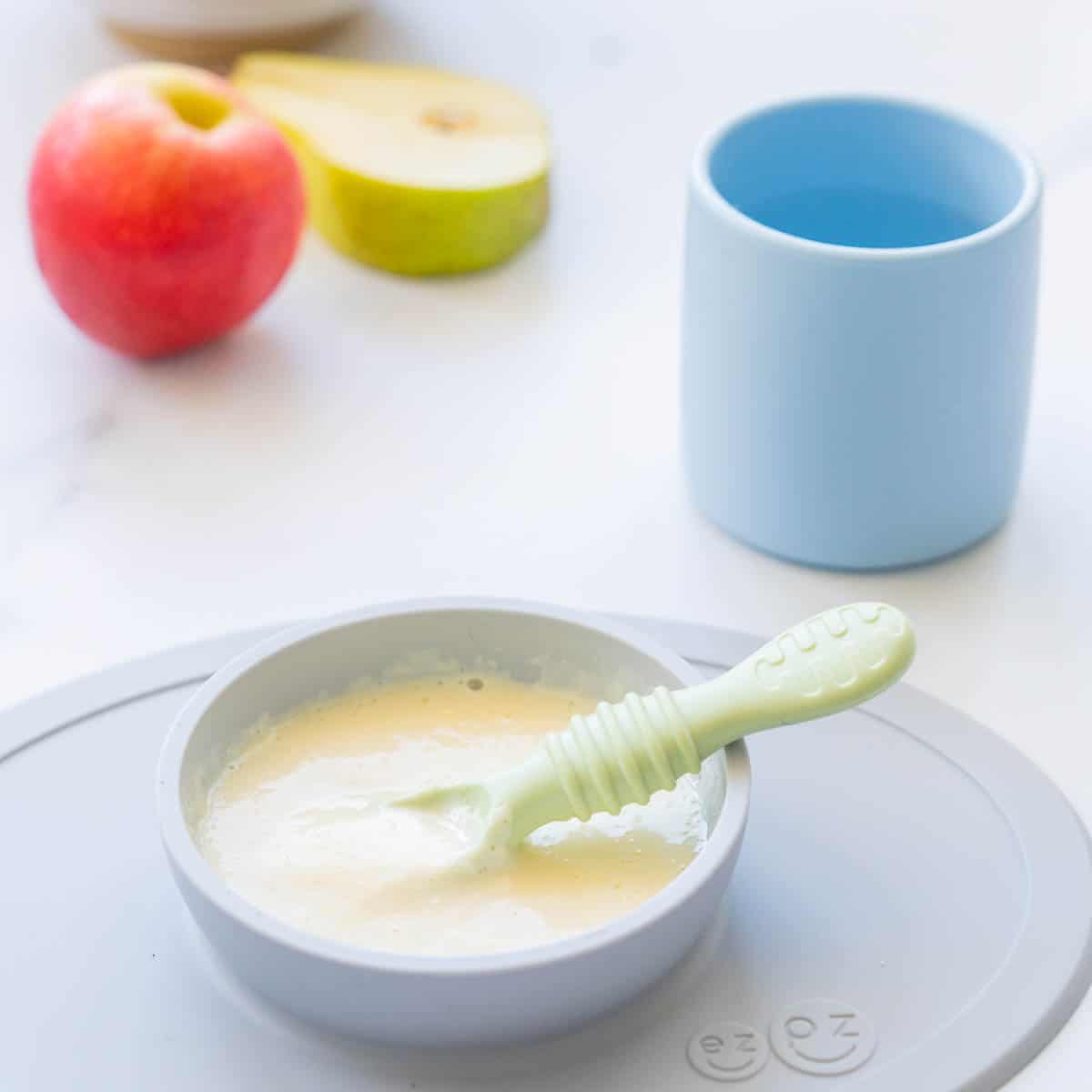 A grey silicone baby plate of baby custard with a green spoon, blue baby cup and fruit in the background.