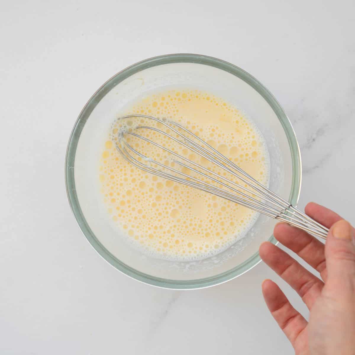 Uncooked egg and milk mixture in a glass mixing bowl with a whisk.