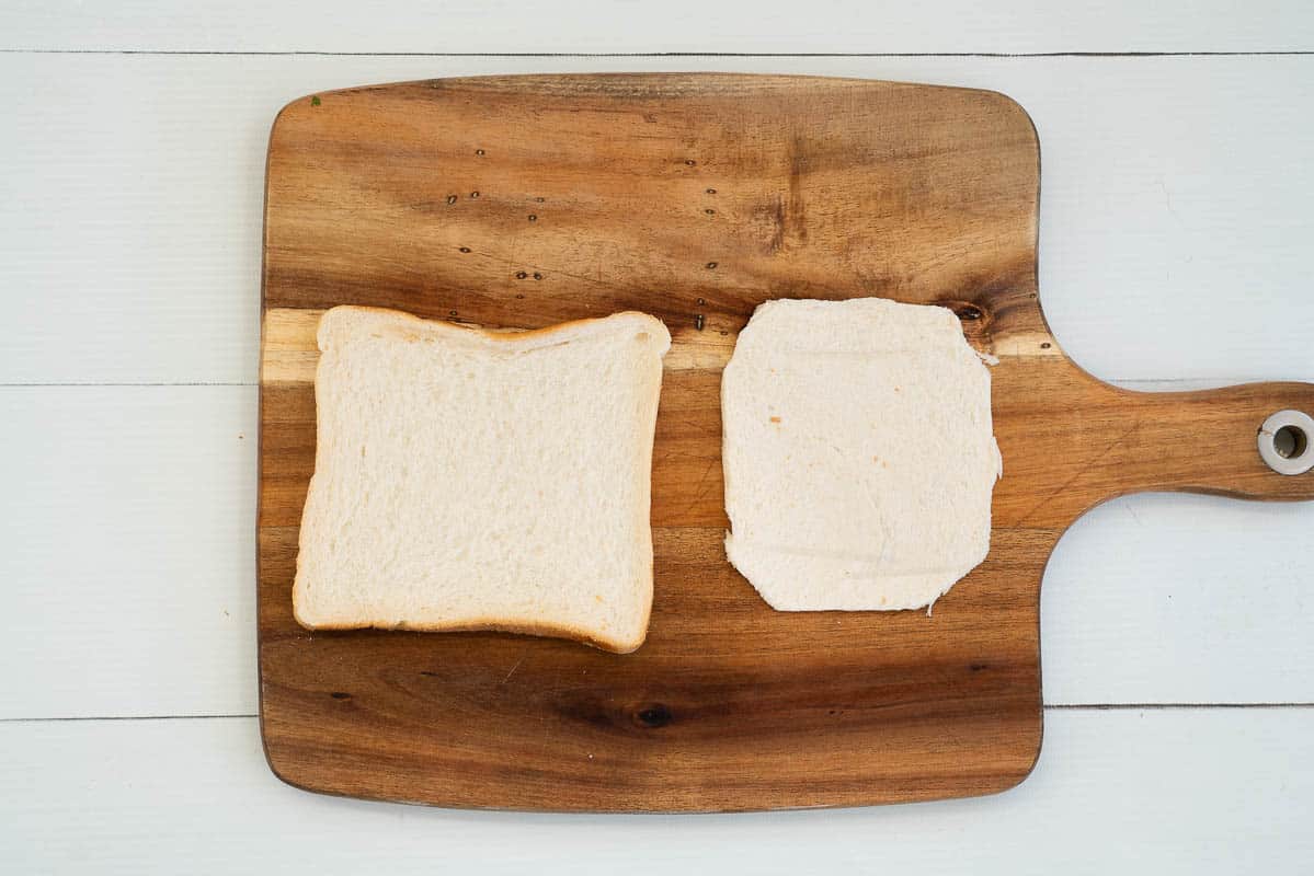 chopping board with 2 slices of bread, one has had the crusts removed and the corners trimmed