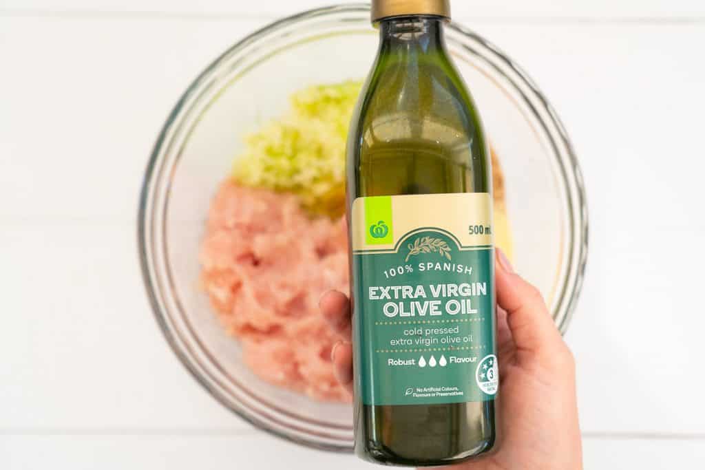 Woman's hand holding a bottle of extra virgin olive oil above a bowl with the chicken rissole ingredients