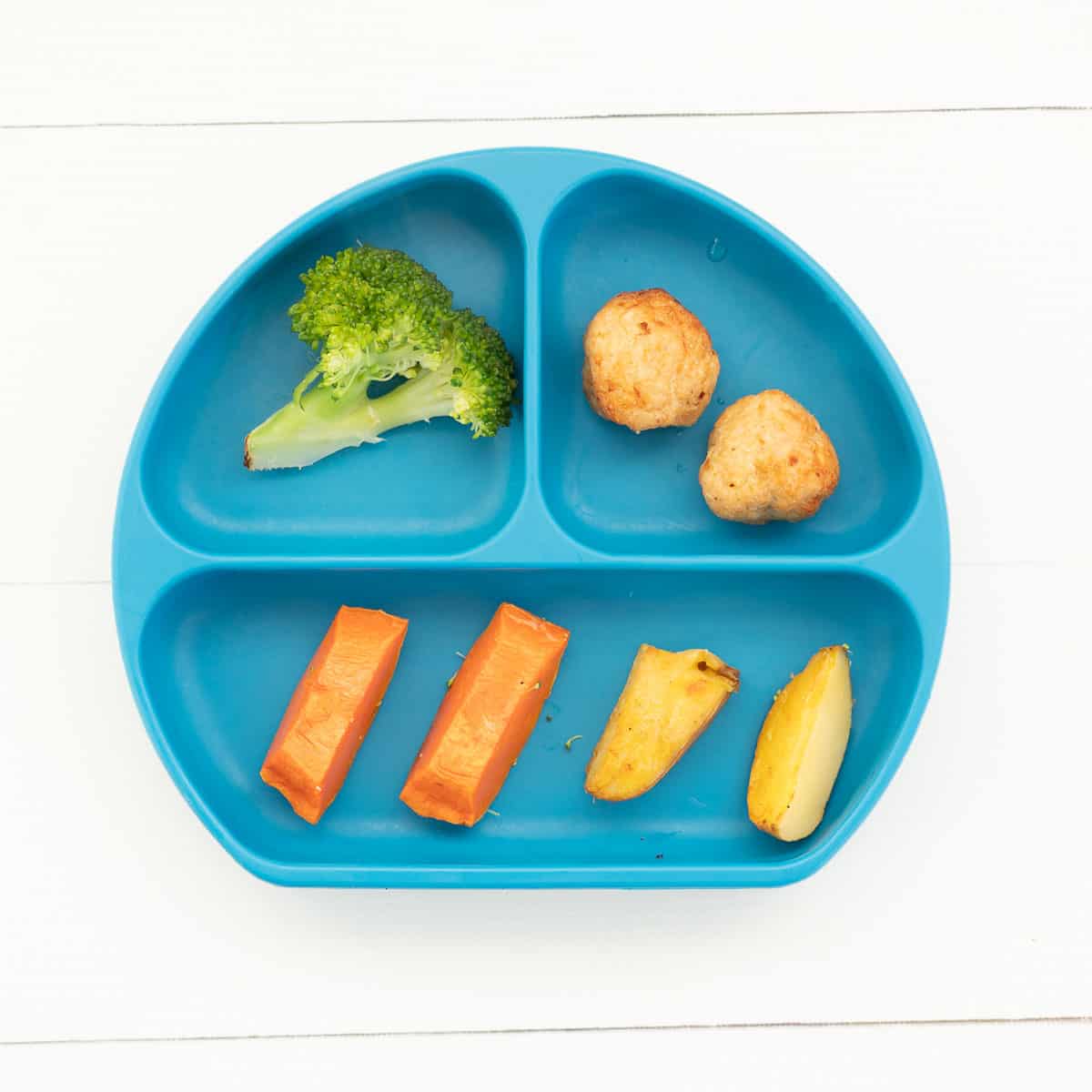 blue silicone divided plate with cooked pieces of pumpkin, potato, broccoli and 2 baked chicken meatballs