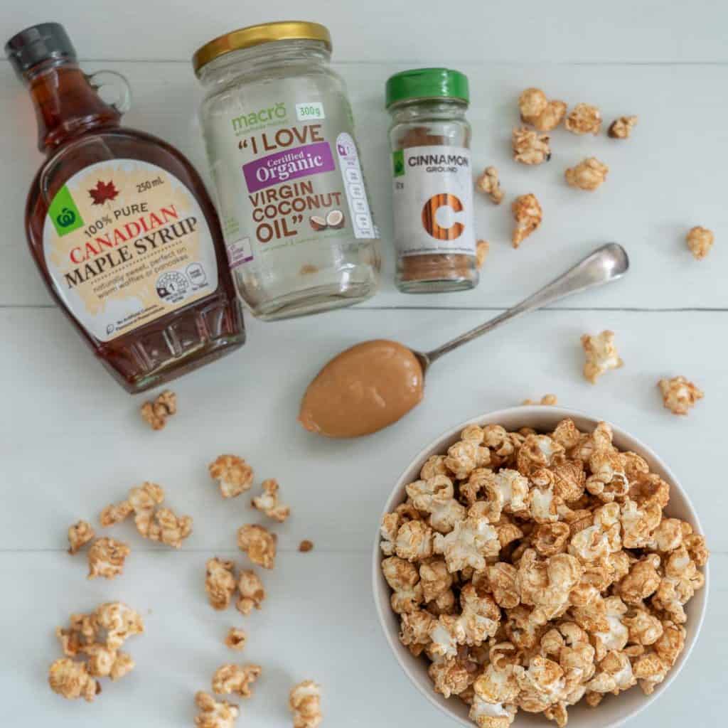 a bowl of cinnamon popcorn with the ingredients for the recipe maple syrup, coconut oil, cinnamon and nut butter