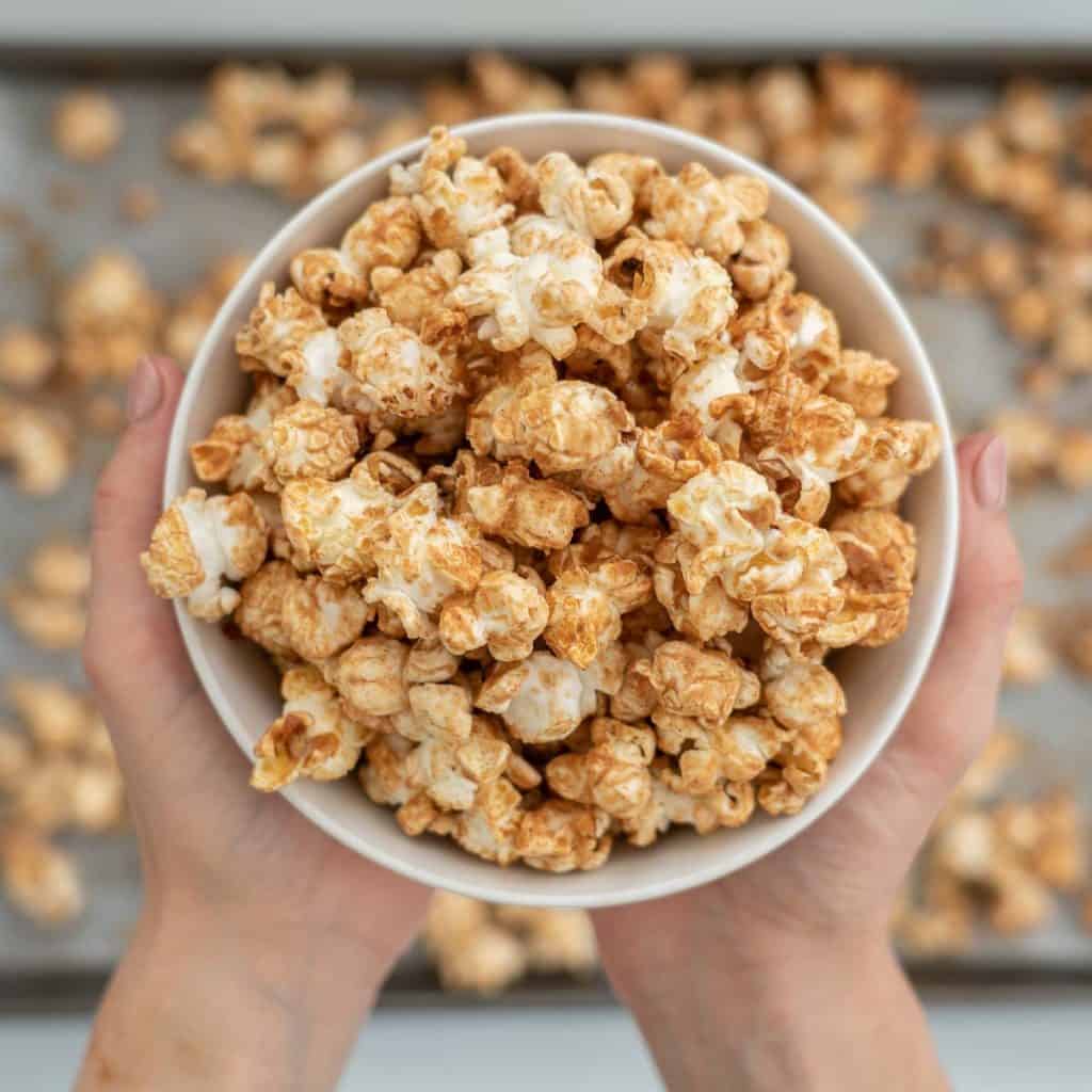 two hands holding a bowl of caramel coloured popcorn above a baking tray covered in popcorn