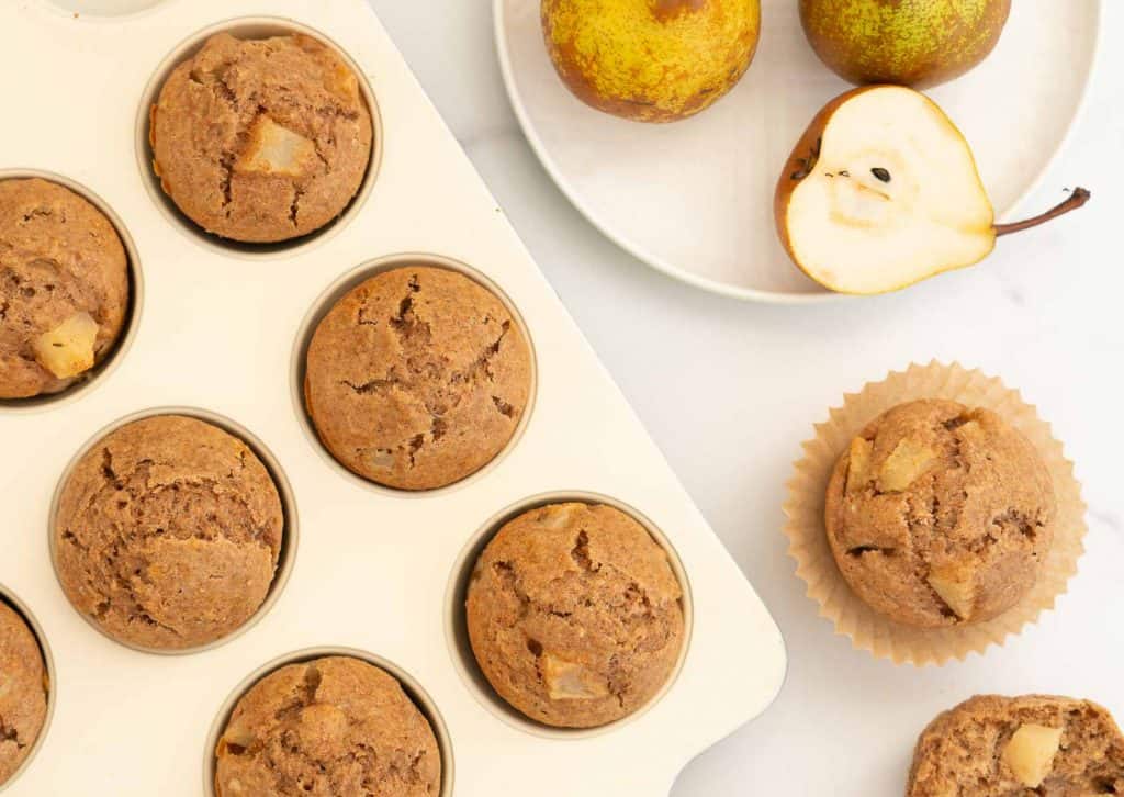 Top view of muffins in a cream muffin tin, chunks of pear visible in the top of the muffins, a plate of pears sitting on the marble bench top next to the muffin tin