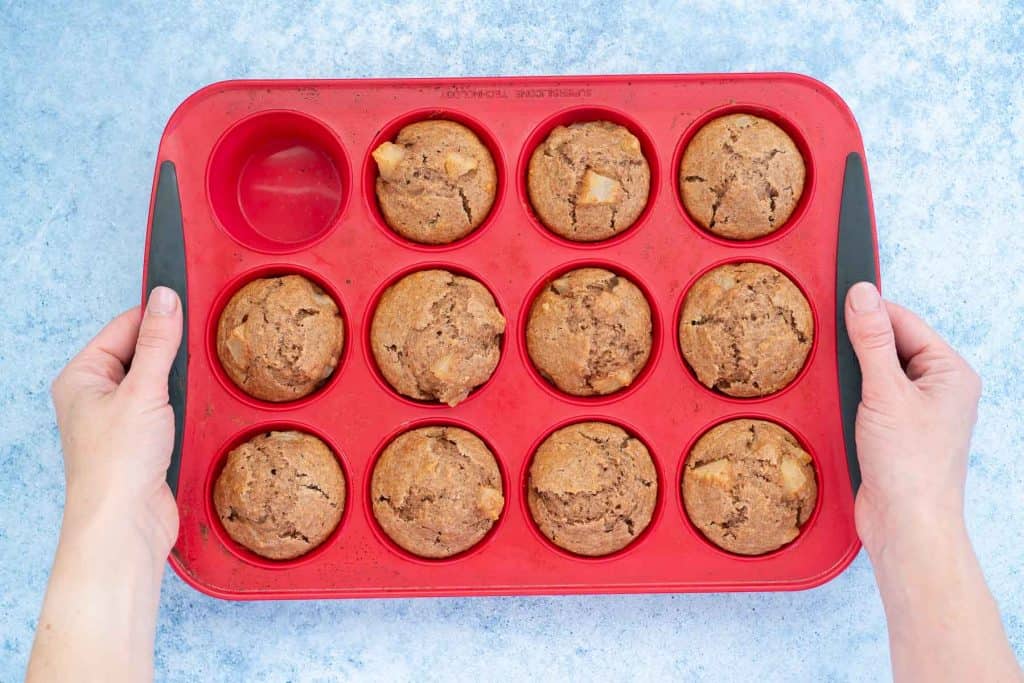 11 cooked muffins in a 12 cup red silicone muffin tray, tray held on either side by a woman's hands