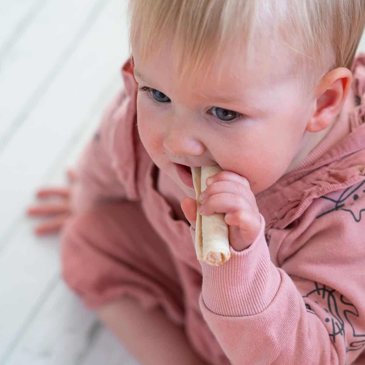 1 year old baby sitting on the floor chewing on a teething rusk