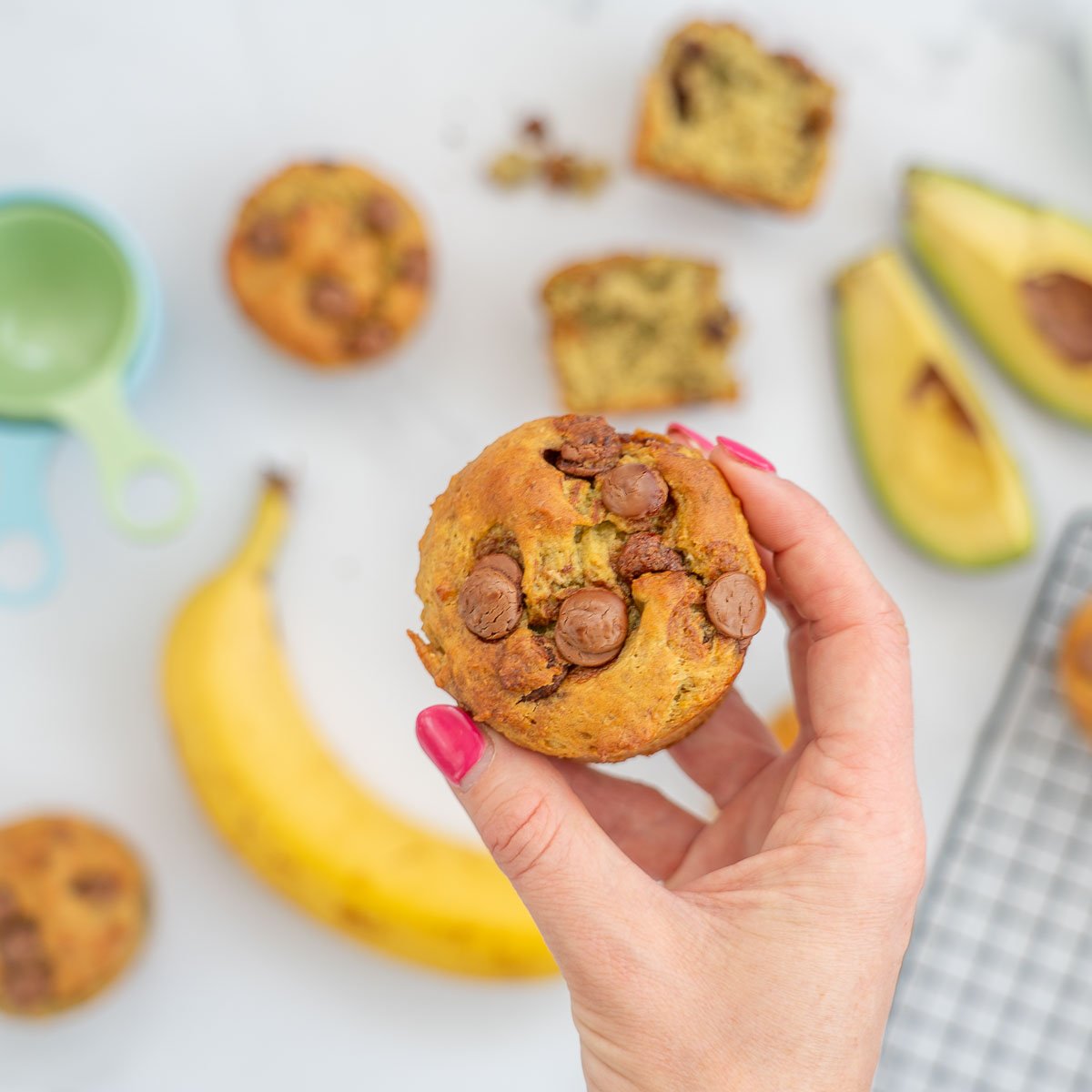 Woman's hand holding a muffin above a bench top covered with muffins, banana and avocado