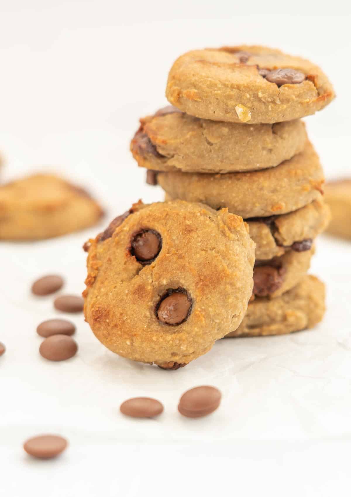 A stack of 6 cookies with one cookie leaning against the stack, chocolate ships scattered around the base