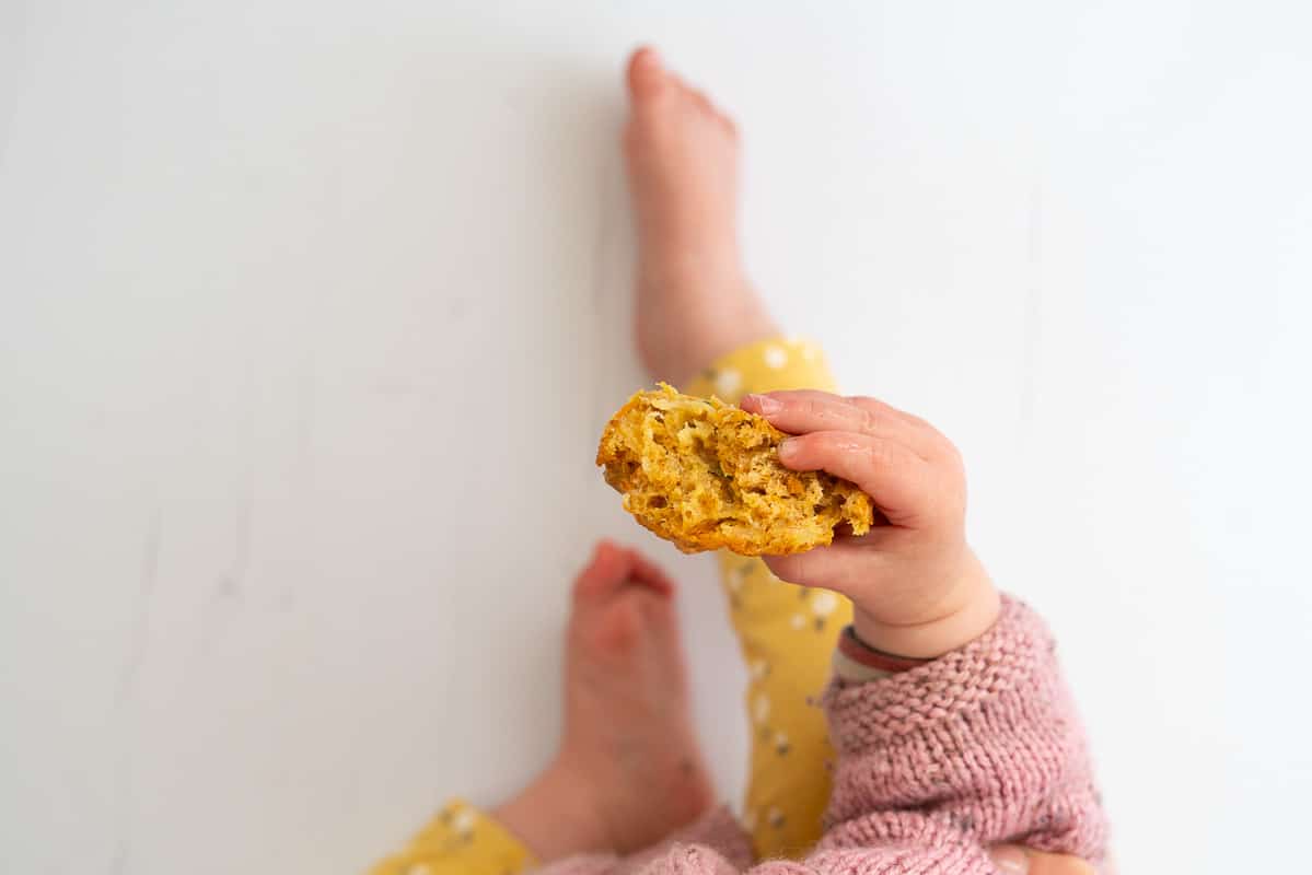 baby sitting on a floor holding a half eaten muffins