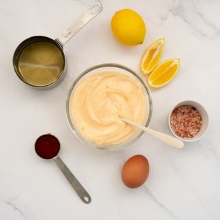 glass bowl of sriracha aioli surrounded by ingredients (oil, lemon, sriracha, salt, egg)